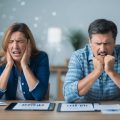 A husband and wife real estate investing team realizing they have become victims of title fraud. The wife is crying and throwing something and the husband is clenching his fists in after.