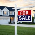 A house with a "For Sale" sign in front of it, surrounded by similar houses in a suburban neighborhood