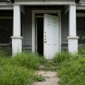 A vacant house with the front door open, overgrown grass and neglected exterior, a sense of abandonment and potential investment.