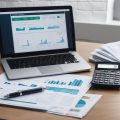 An open laptop displaying real estate CMA charts and graphs on a desk with a stack of papers and a calculator