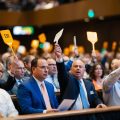 Bidders jostle, waving paddles, at a crowded foreclosure auction. Auctioneer calls out prices as tension mounts