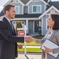 A real estate agent shaking hands with a homeowner in front of a suburban house with a for sale sign.