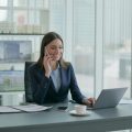 A real estate operations assistant working at a desk in a modern office with a laptop and paperwork, smiling confidently.