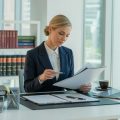 A real estate paralegal reviewing legal documents at a desk in a modern office with shelves of books and a city view outside the window.