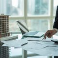 A professional reviewing blueprints and financial documents with a building model on a table in a modern office with city views.