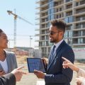 Two business professionals discussing real estate plans near a new residential building under construction.