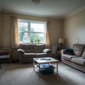 Interior of a modest living room showing signs of wear and tear, with basic furniture and natural light coming through a window.