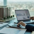 A business professional reviewing financial documents and real estate models in an office with a city view.