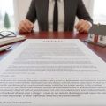 Close-up of a real estate deed document on a desk with reading glasses, a pen, house keys, and a small house model nearby.