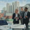 Business professionals discussing real estate plans outdoors with city buildings and shipping containers visible in the background.