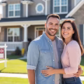 A smiling military veteran couple standing in front of their new suburban home with a lawn and porch on a sunny day.