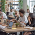 A modern co-working space with people working together at desks and using laptops in a bright room with large windows and plants.