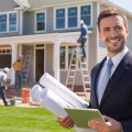 A real estate investor holding blueprints and a tablet stands outside a partially renovated house with construction workers actively working in the background.