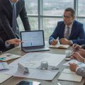 A group of business professionals reviewing documents and a laptop in an office during a real estate investment meeting.