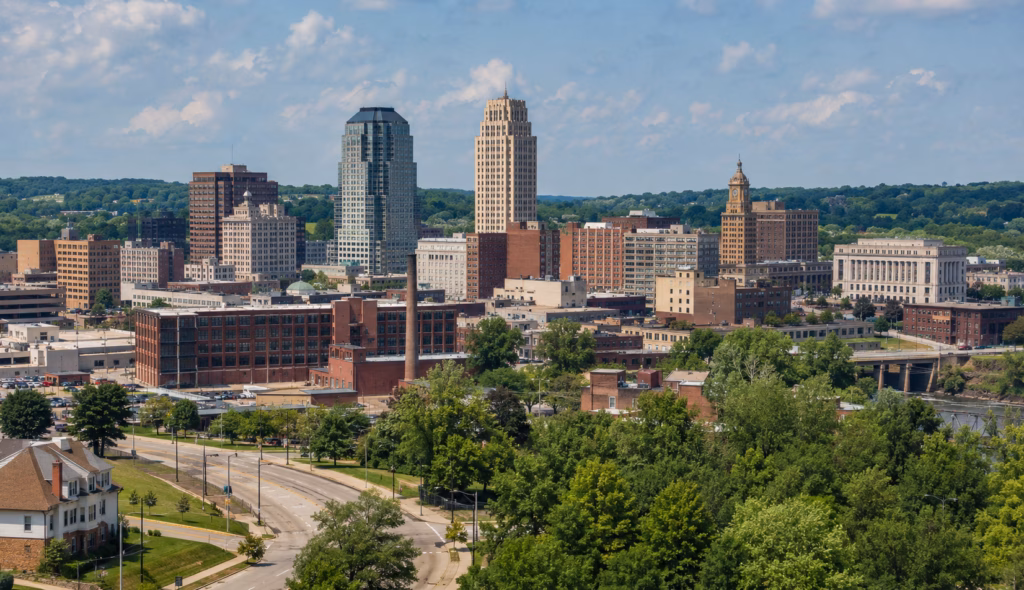 Urban skyline with mid-rise office towers, industrial buildings, leafy neighborhoods, and rolling green hills in the distance.