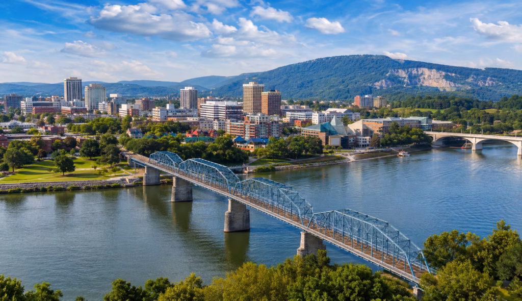 Riverfront cityscape with downtown buildings, bridges over the Tennessee River, green hills, and Lookout Mountain in the background.