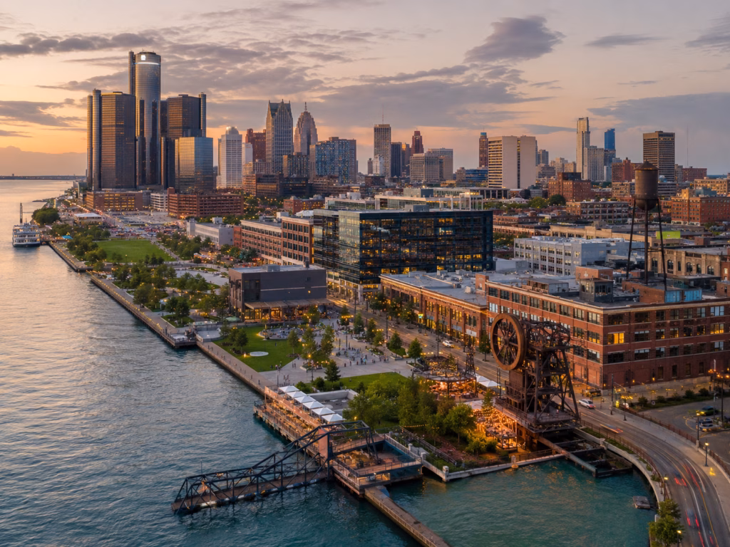 Detroit waterfront skyline at sunset with the riverfront promenade, industrial heritage elements, and downtown high-rises.