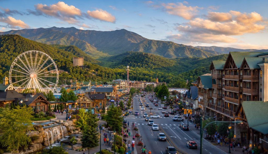 Mountain resort town with the Great Smoky Mountains, chalet-style buildings, tourist attractions, a Ferris wheel, and a busy main road at sunset.