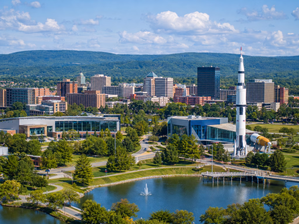 Huntsville skyline with a lakeside park, fountain, and rocket exhibit set against green hills and modern buildings.