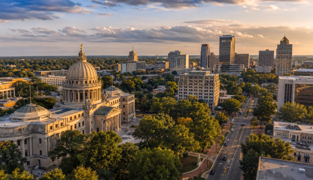 Downtown Jackson skyline with the domed state capitol, office towers, broad streets, mature trees, and warm sunset light.