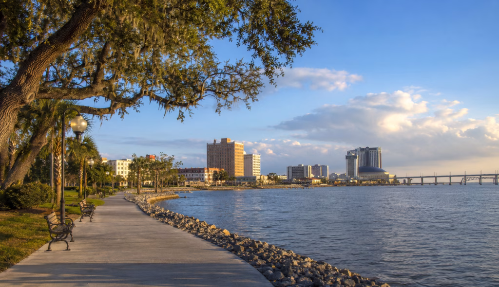 Waterfront promenade with live oak trees, benches, lake views, bridge, and downtown buildings at golden hour.