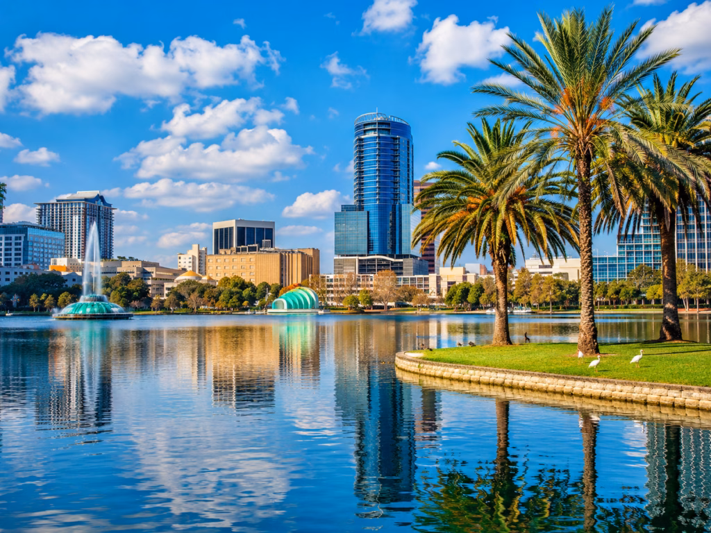Orlando skyline reflected across a lake with palm trees, fountains, and sunny waterfront park scenery.