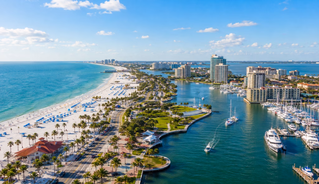 Aerial view of white-sand beaches, turquoise coastal water, palm-lined roads, marinas, boats, and high-rise waterfront buildings.