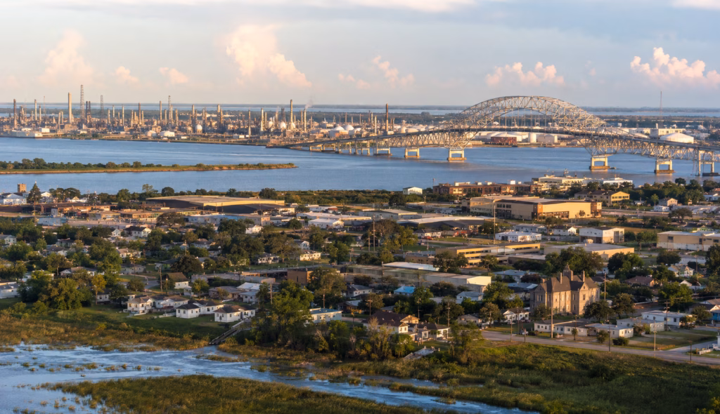 Waterfront industrial cityscape with refineries, shipping channels, marshland, and a large arched bridge along the Gulf Coast.